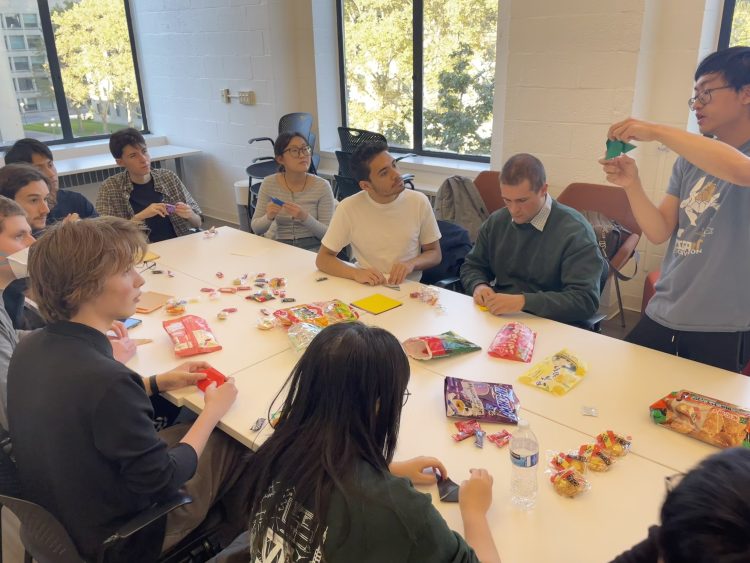 ten people sitting around a large table with snacks and origami paper, while one student demonstrates origami technique